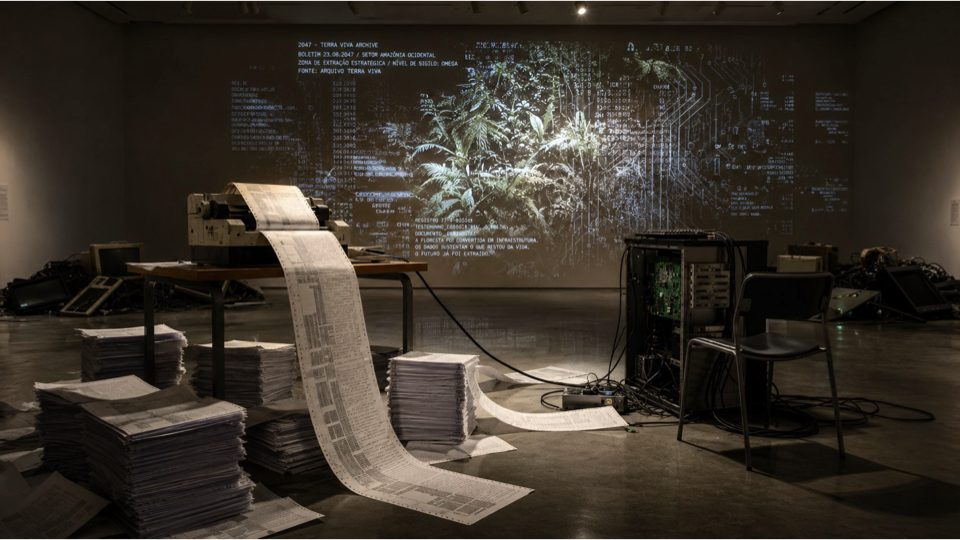 Installation view with a dot-matrix printer, strips and stacks of paper, an open computer and a wall projection